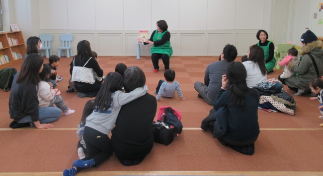 親の膝に乗ったり抱き着いたりしながら読み聞かせを聞く子どもたちの写真