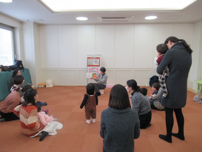 絵本を読む先生とそれを見る子供とお母さんの写真
