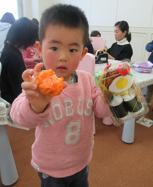 詰めたお弁当とオレンジ色の紙でできたみかんを見せる子どもの写真