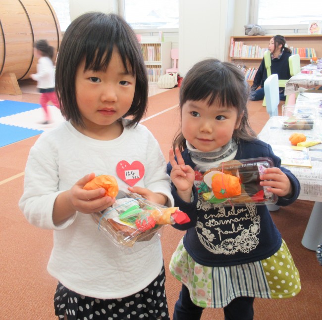 詰めたお弁当とみかんを持ってポーズをとる二人の女の子の写真