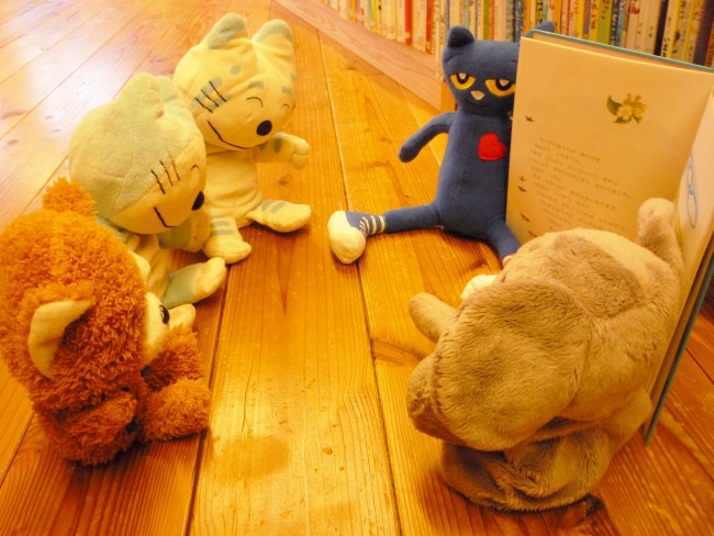 ゾウとスニーカーを履いた猫のぬいぐるみが読み聞かせをし、ほかのぬいぐるみがそれを聞いている風の写真