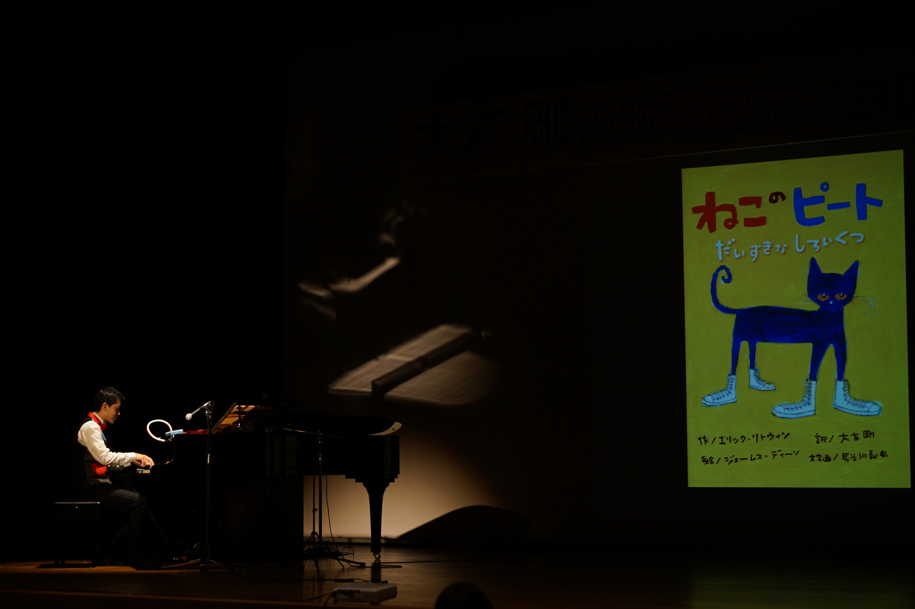 スクリーンに映された絵本「猫のピート」の表紙の画像の前でピアノを弾く大友さんの写真