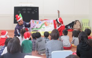 サンタの扮装をしたスタッフが子どもたちに絵を見せながら読み聞かせをしている写真